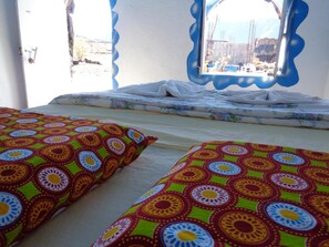 A room with two beds, each with colorful patterned pillows, a window with a blue frame, and a view of a construction site outside. - Pensao Casa José Doce (Santa Catarina do Fogo)