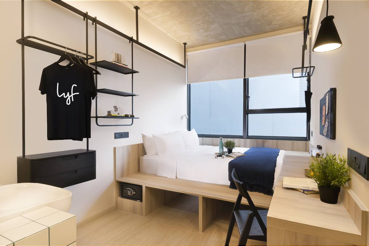 A compact bedroom with a bed, bedside table, and a hanging black t-shirt with 'lyf' written on it.