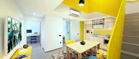 A modern living room with a yellow accent wall, a dining area with a patterned rug, and a kitchen with wooden cabinets.