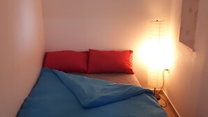 A small, narrow room with a bed, a red pillow, a blue blanket, and a wall-mounted lamp. - I'm Jerusalem (Jerusalem)