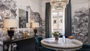 A dining area with a round table, two chairs, a chandelier, and a view of buildings through a window. - Rocco Forte House Piazza di Spagna Roma (Rome)
