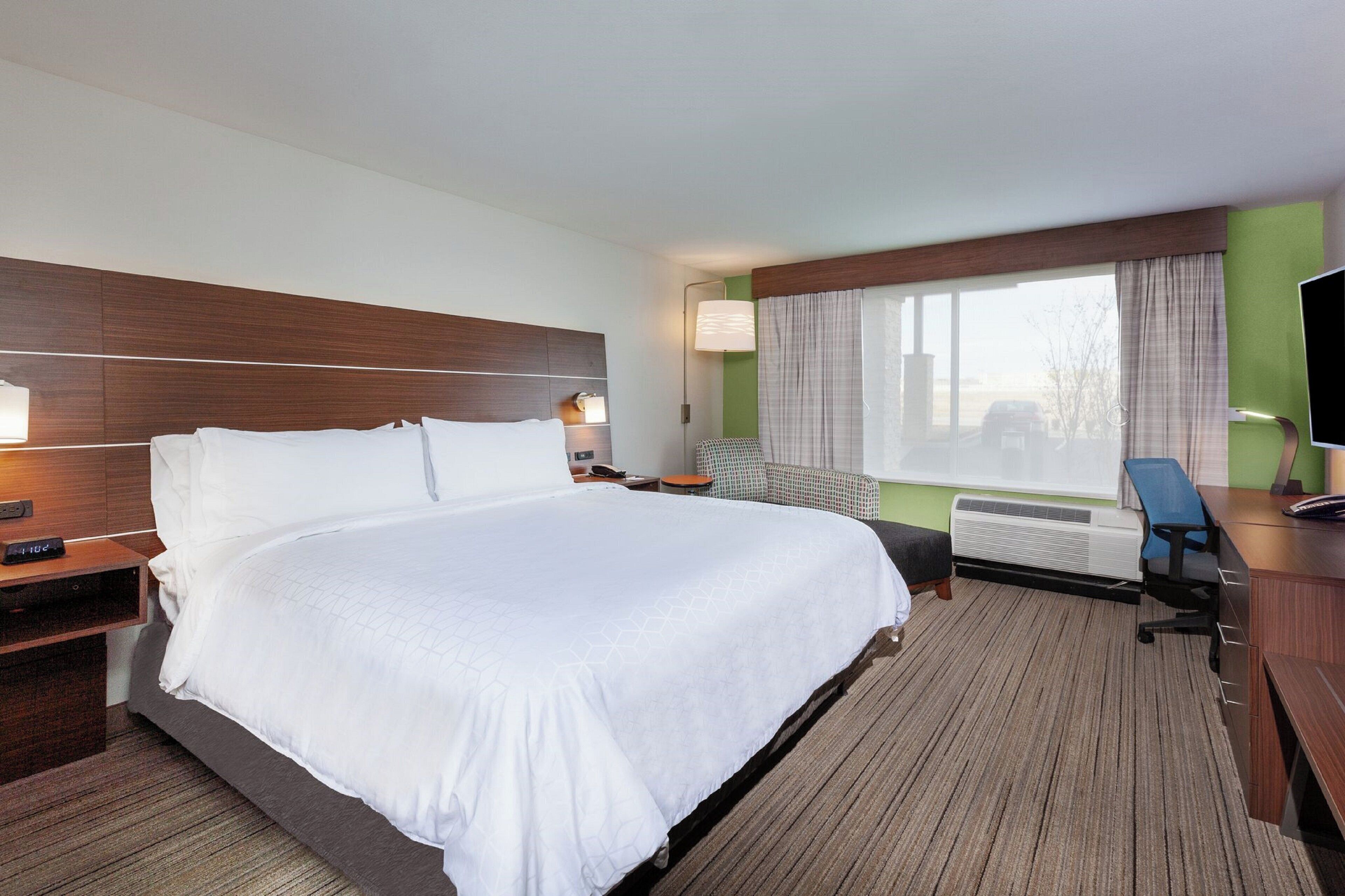 Quarto de hotel com uma cama grande, uma escrivaninha, uma cadeira, uma televisão e uma janela com cortinas.