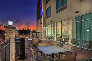 An outdoor seating area with tables and chairs, a trash can, and a building with large windows. - Holiday Inn Express & Suites Tulsa Northeast - Owasso by IHG (Owasso)