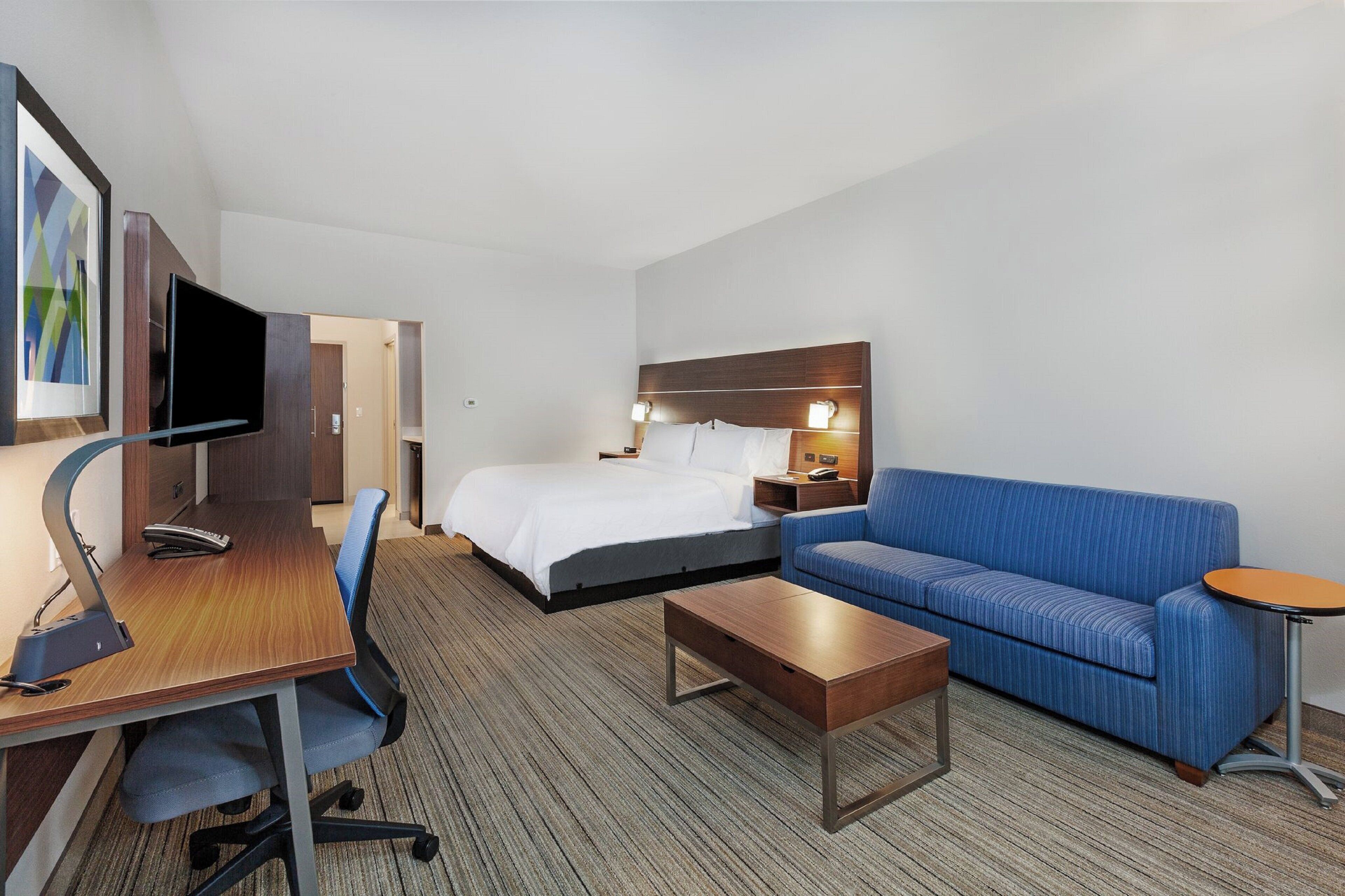 Een hotelkamer met een bed, een bureau met een computer, een blauwe bank, een klein tafeltje en een aan de muur bevestigde televisie.