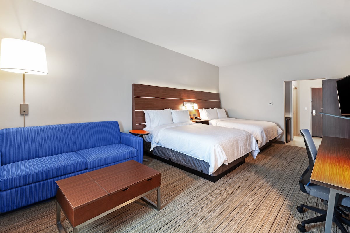 A hotel room with a blue sofa, a wooden coffee table, a bed with two pillows, a nightstand, and a desk with a chair.