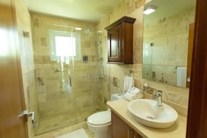 A bathroom with a glass-enclosed shower, a white toilet, a sink with a mirror above it, and a wooden cabinet. - Escondido Bay Properties by Caribe Stays (Sosúa)