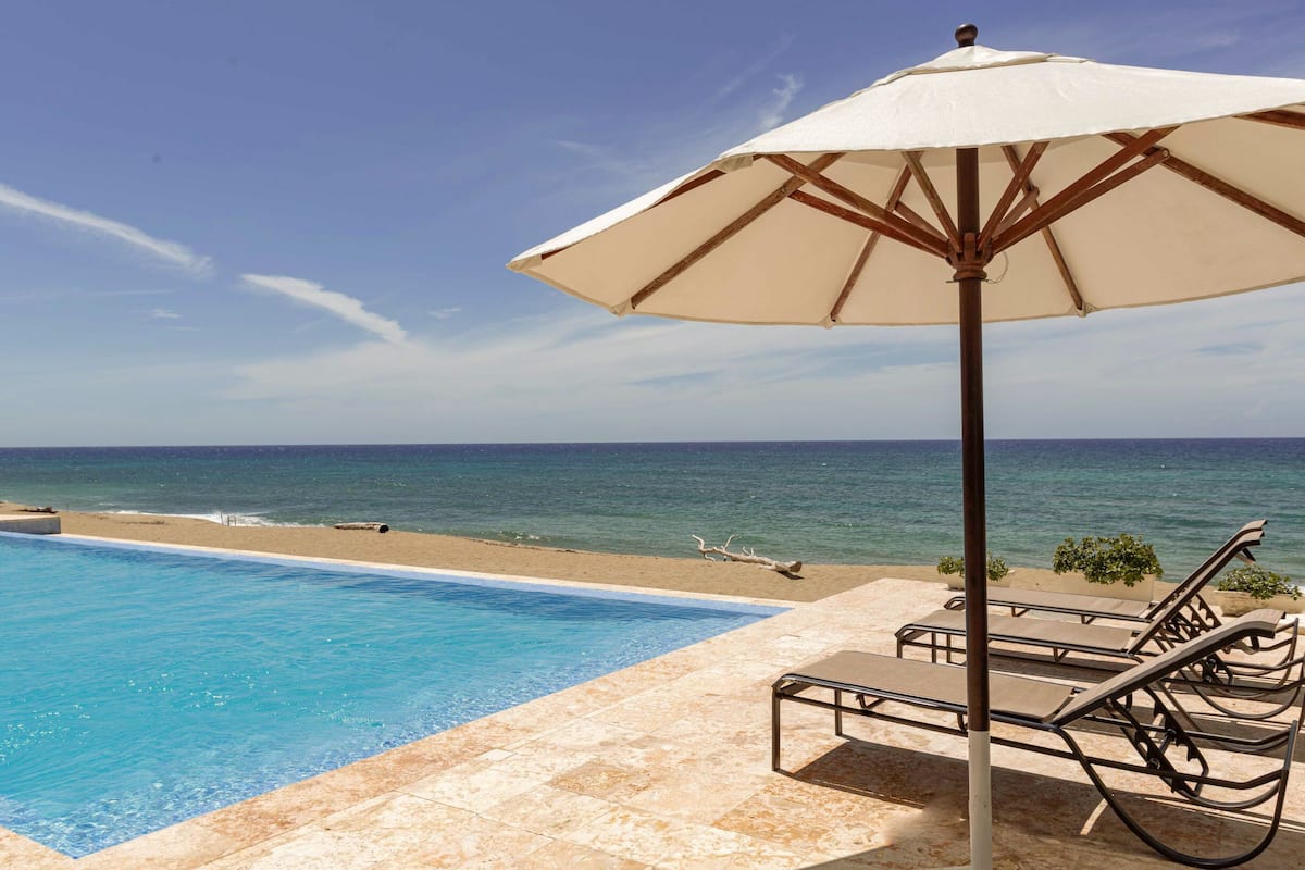 A poolside area with a view of the ocean, lounge chairs, and an umbrella.