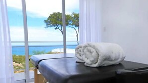 Een massagetafel met een handdoek erop, uitzicht op zee en bomen door grote ramen.