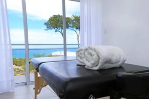 Uma mesa de massagem com uma toalha, vista para o mar e árvores através de grandes janelas.
