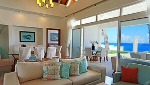 A spacious living area with a large sofa, a smaller armchair, and a dining table. There are multiple chairs around the table, and a large window offers a view of the ocean. - Ocean Front Villa Deluxe 4 Bedroom (Sosúa)