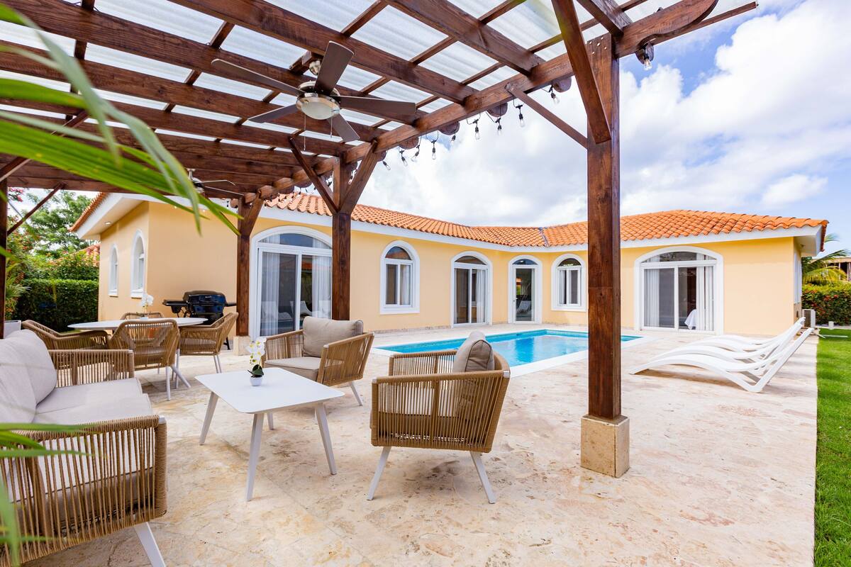 A covered outdoor patio with wicker furniture, a pool, and a house with a tiled roof.