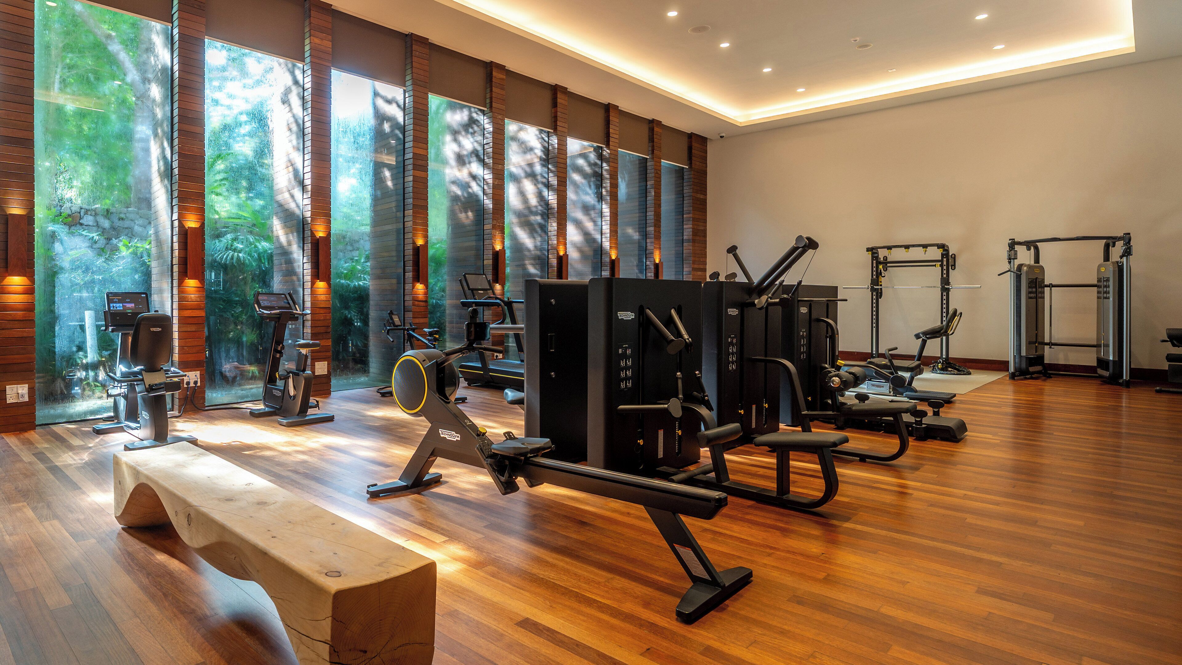 A gym with exercise equipment, large windows, and a wooden bench.