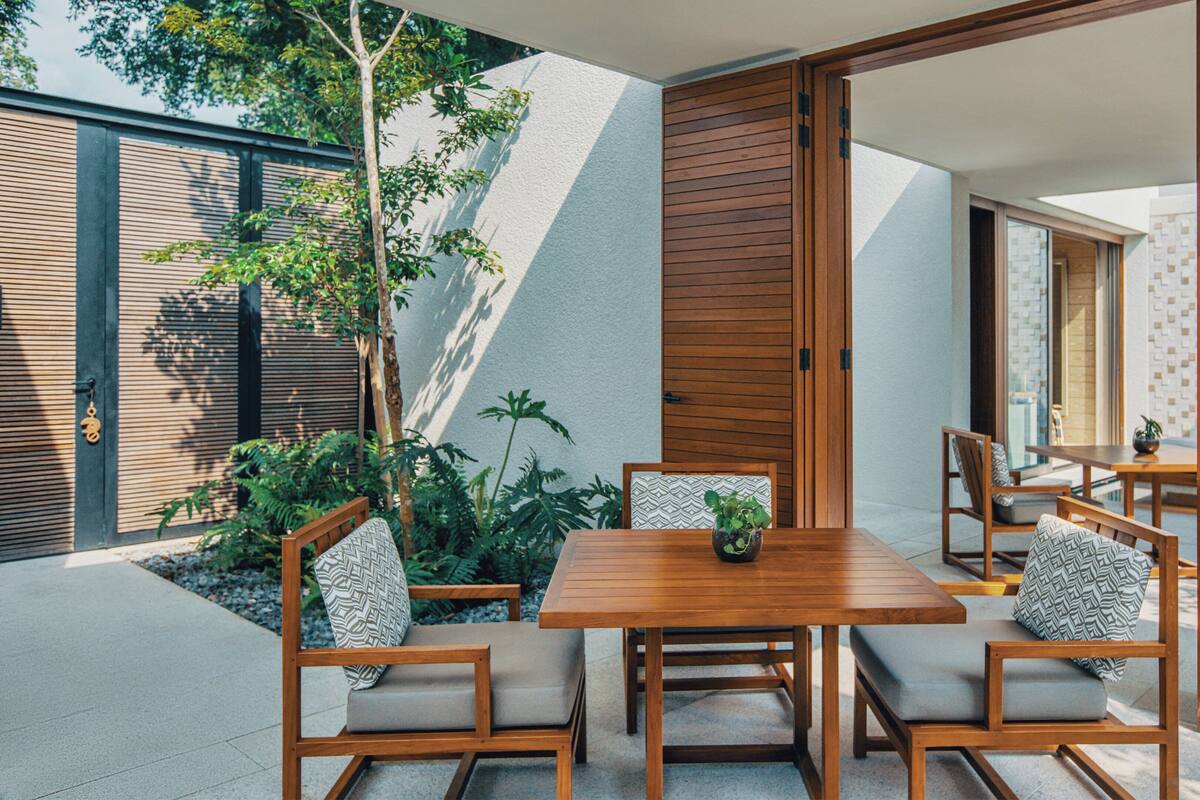 An outdoor dining area with a wooden table and chairs, surrounded by plants and a modern architectural design.