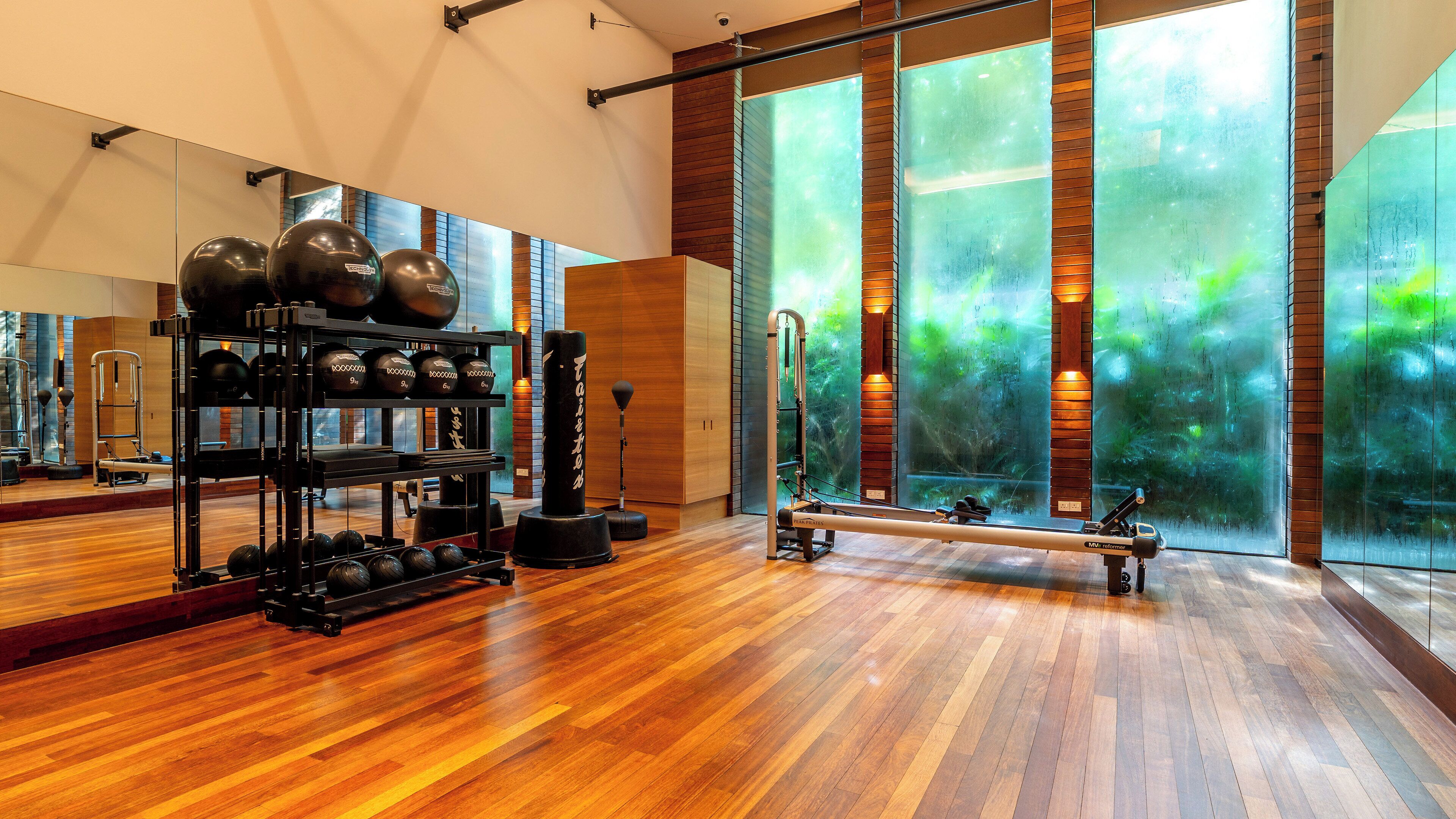 Un gimnasio amplio con suelo de madera, equipamiento de ejercicio y grandes ventanales con vista a zonas verdes.