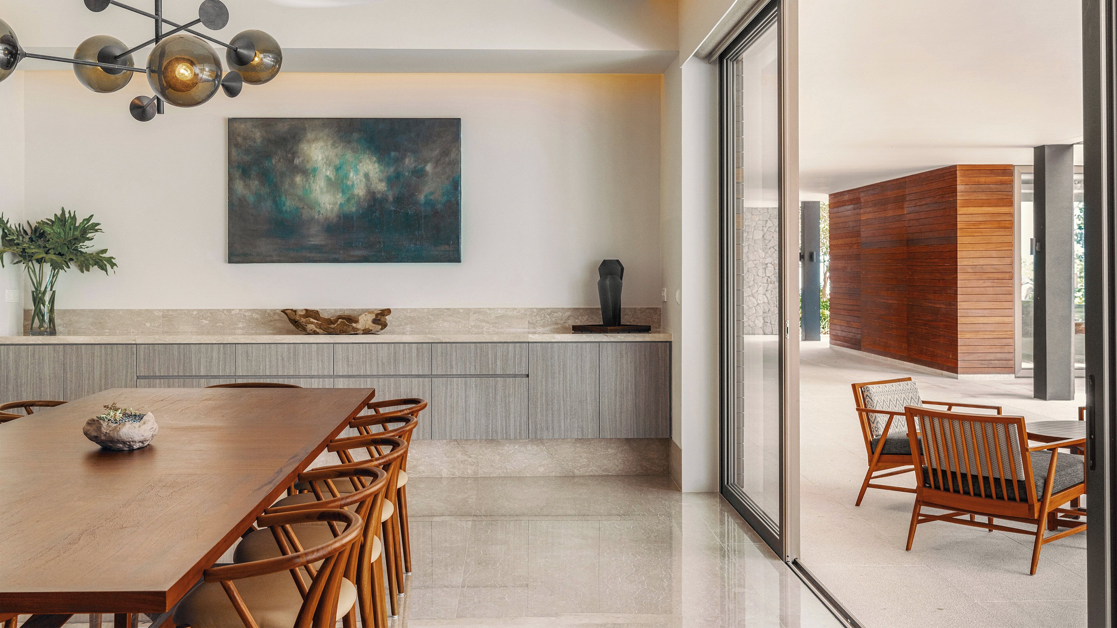 A modern dining area with a wooden table and chairs, a large abstract painting on the wall, and a view of the outdoors through a glass door.