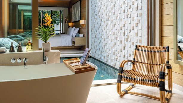 A modern bathroom with a freestanding bathtub, a wooden chair, and a large mirror reflecting a bedroom with a bed and a view of a pool. - Mandarin Oriental, Desaru Coast (Bandar Penawar)