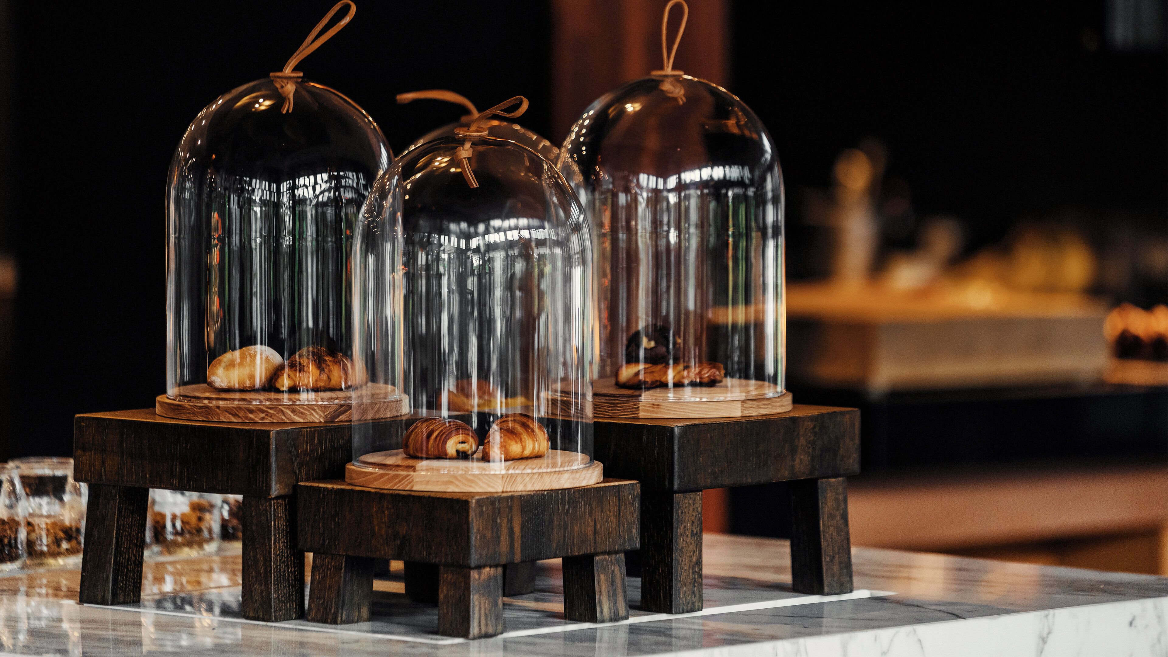 Three glass domes with pastries inside, placed on wooden stands on a marble surface.
