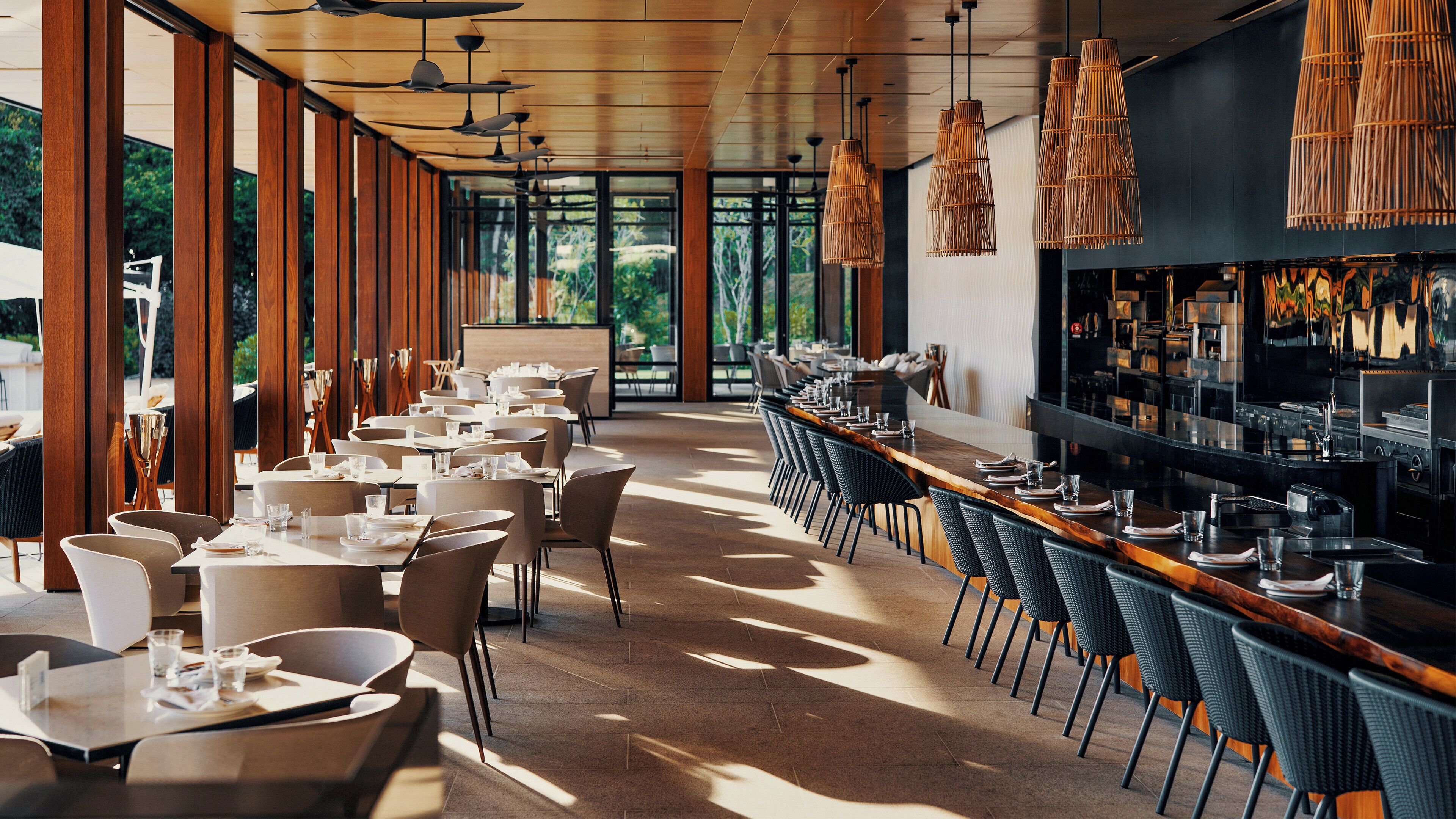 Un restaurante moderno con mesas y sillas largas, ventanas amplias y lámparas colgantes de bambú.