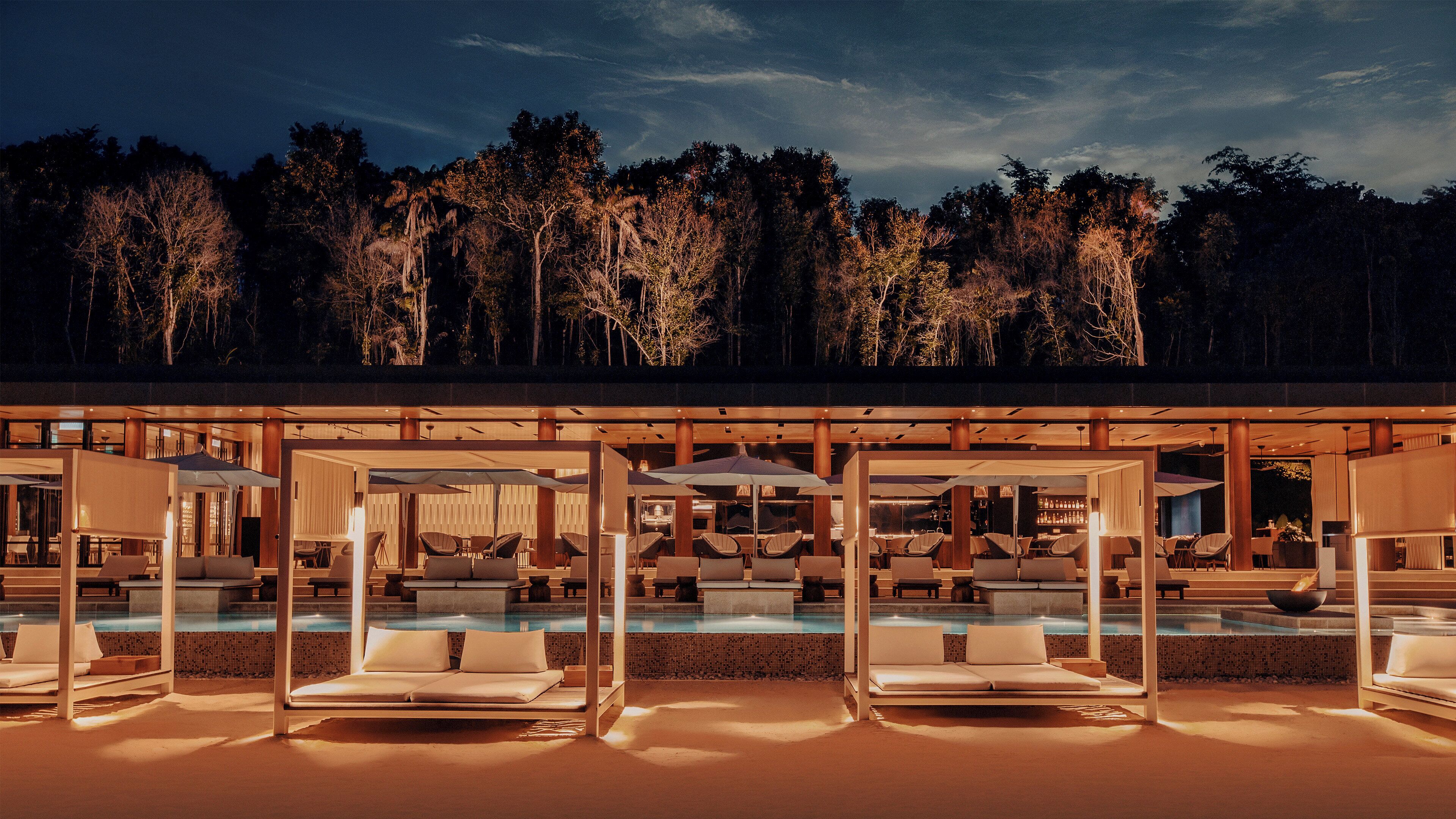 A poolside area with lounge chairs and umbrellas, a bar, and a view of a forested area at dusk.