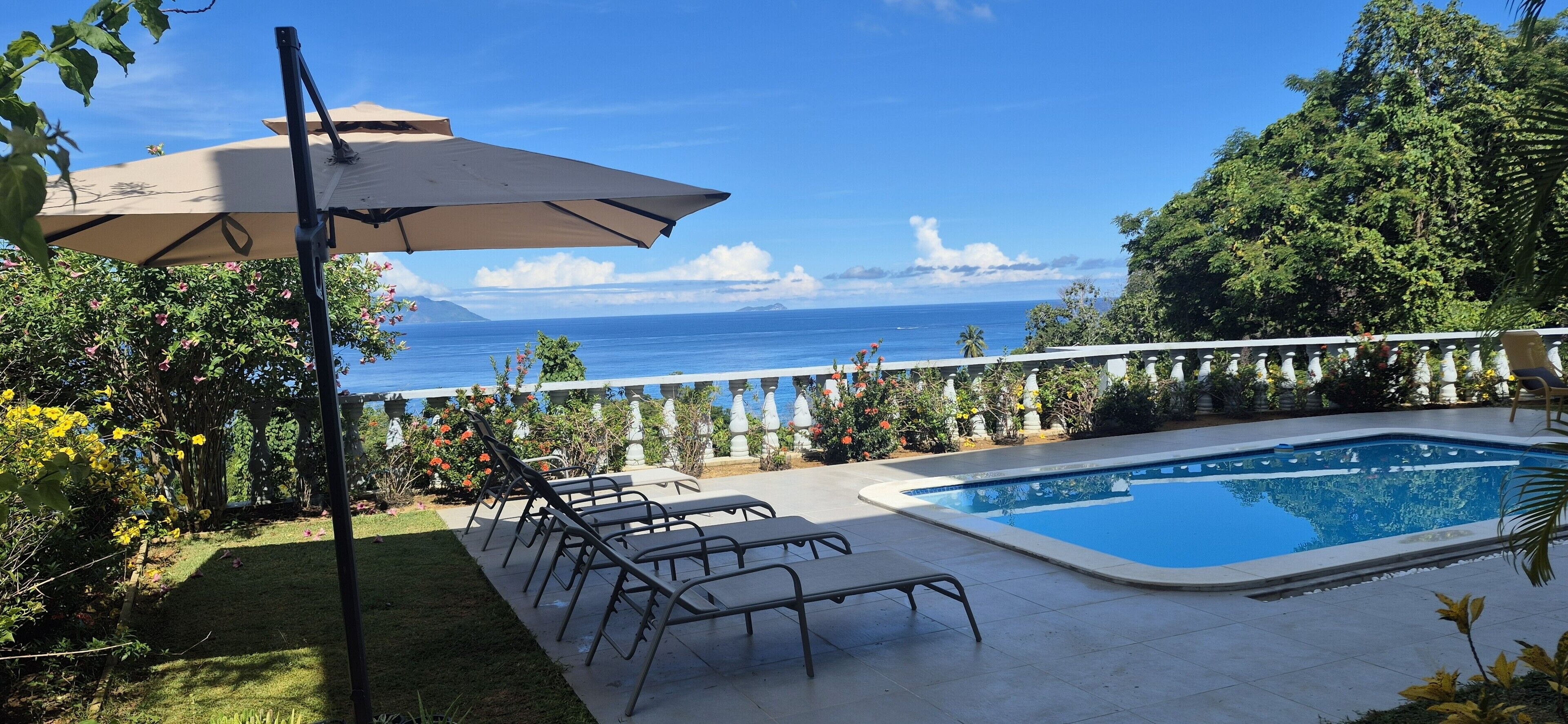 Zona de piscina con sillones de descanso y una sombrilla, rodeada de exuberante vegetación y vistas al océano.