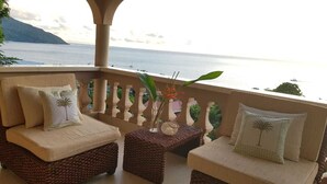 A balcony with two wicker chairs, each with a cushion and a palm tree pillow. A small table with a decorative vase and a candle is between the chairs. The balcony overlooks a body of water with boats and a distant mountain.