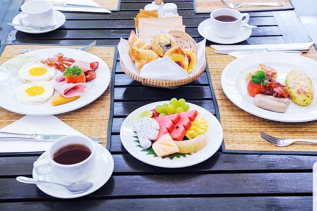 A breakfast setting with plates of eggs, bacon, pastries, and fruit on a wooden table.