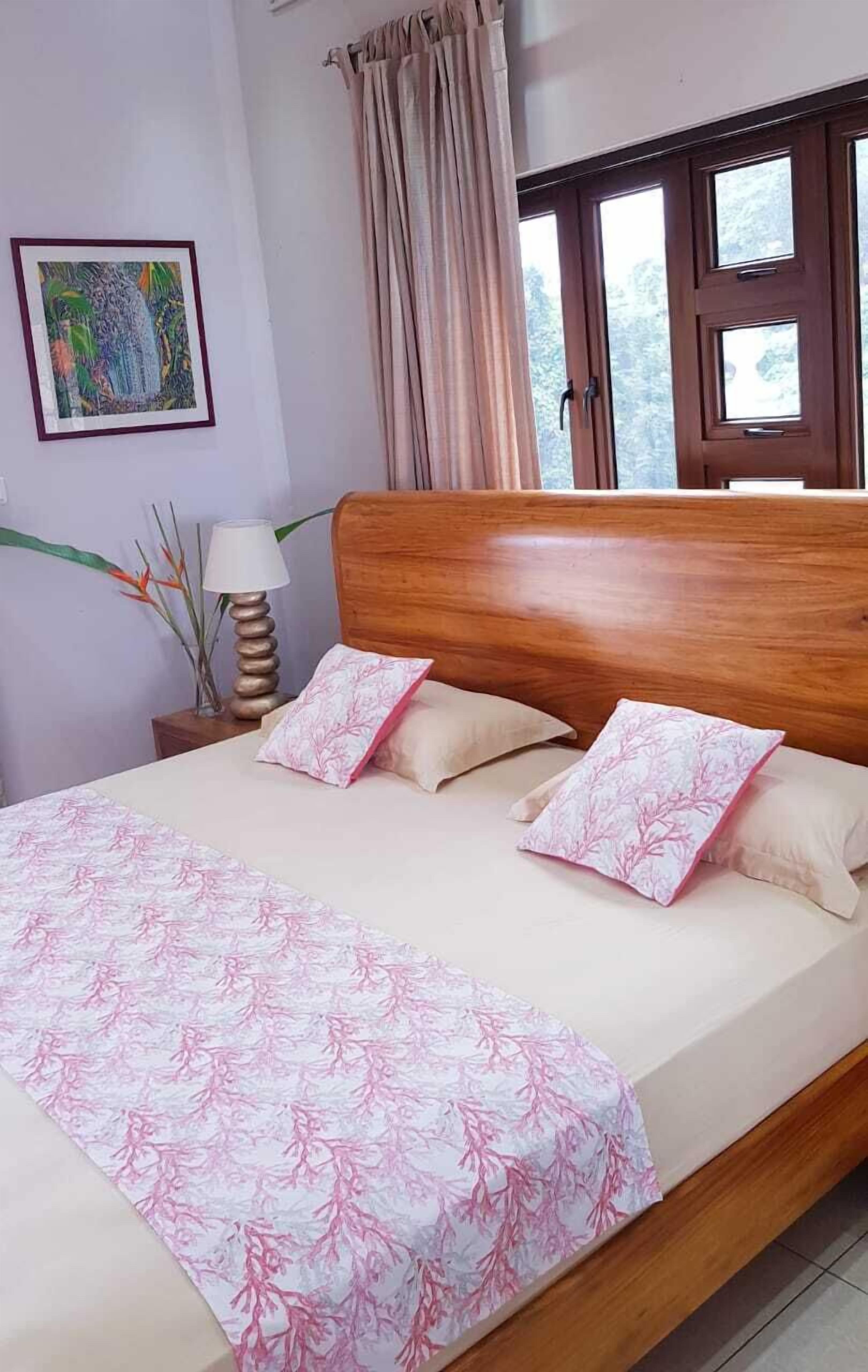 A neatly made bed with a floral bedspread and two pillows, a wooden headboard, a nightstand with a lamp, a framed picture on the wall, and a window with curtains.