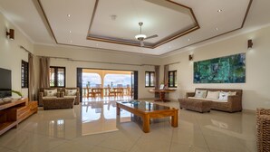 A spacious living room with a large glass coffee table, a ceiling fan, and a large painting on the wall.