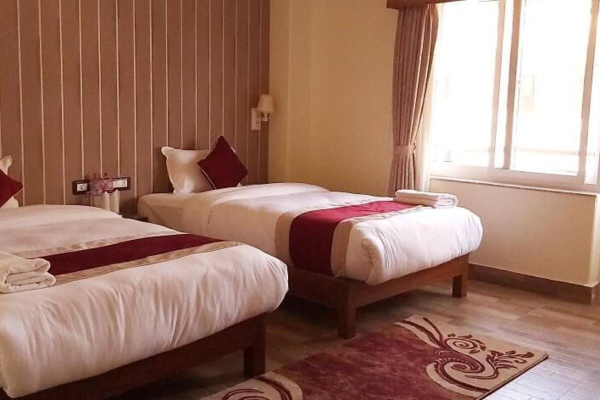 Habitación de hotel con dos camas, una ventana con cortinas, una mesita de noche con lámpara y suelo de madera.