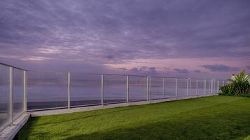 Een balkon met glazen hekwerk, uitzicht op een wateroppervlak, een gazon en een bewolkte lucht.