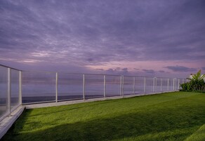 Uma varanda com guarda-corpo de vidro, vista para um corpo de água, área gramada e céu nublado.