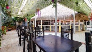 Un restaurant intérieur doté de tables et de chaises à toits de chaume, entouré de plantes suspendues et surmonté d’une structure au plafond de couleur blanche.