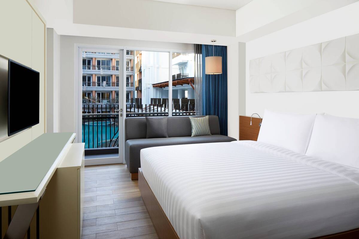 Une chambre d’hôtel avec un lit, un bureau, une télévision, un balcon donnant sur la piscine et une vue sur la ville en arrière-plan.