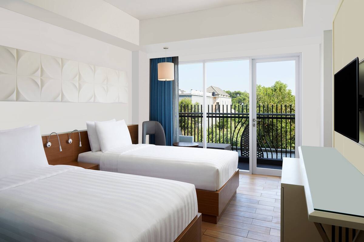 Une chambre d’hôtel avec deux lits, un bureau, une télévision et un balcon offrant une vue sur les arbres et les bâtiments.
