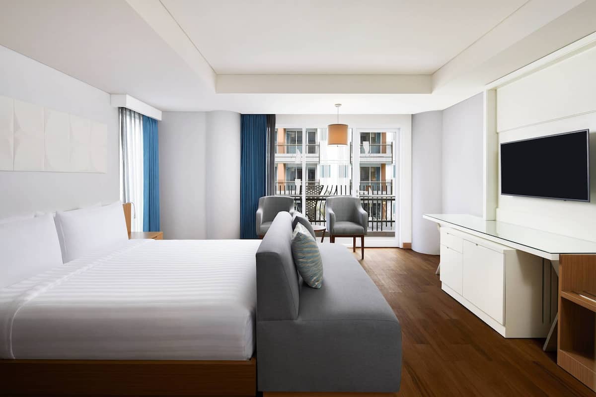 Une chambre d’hôtel moderne avec un grand lit, un canapé gris, une télévision et un balcon avec des chaises.