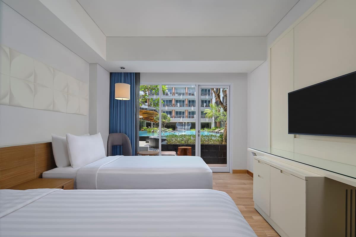 Une chambre d’hôtel avec deux lits, une télévision et une vue sur une piscine et des immeubles.