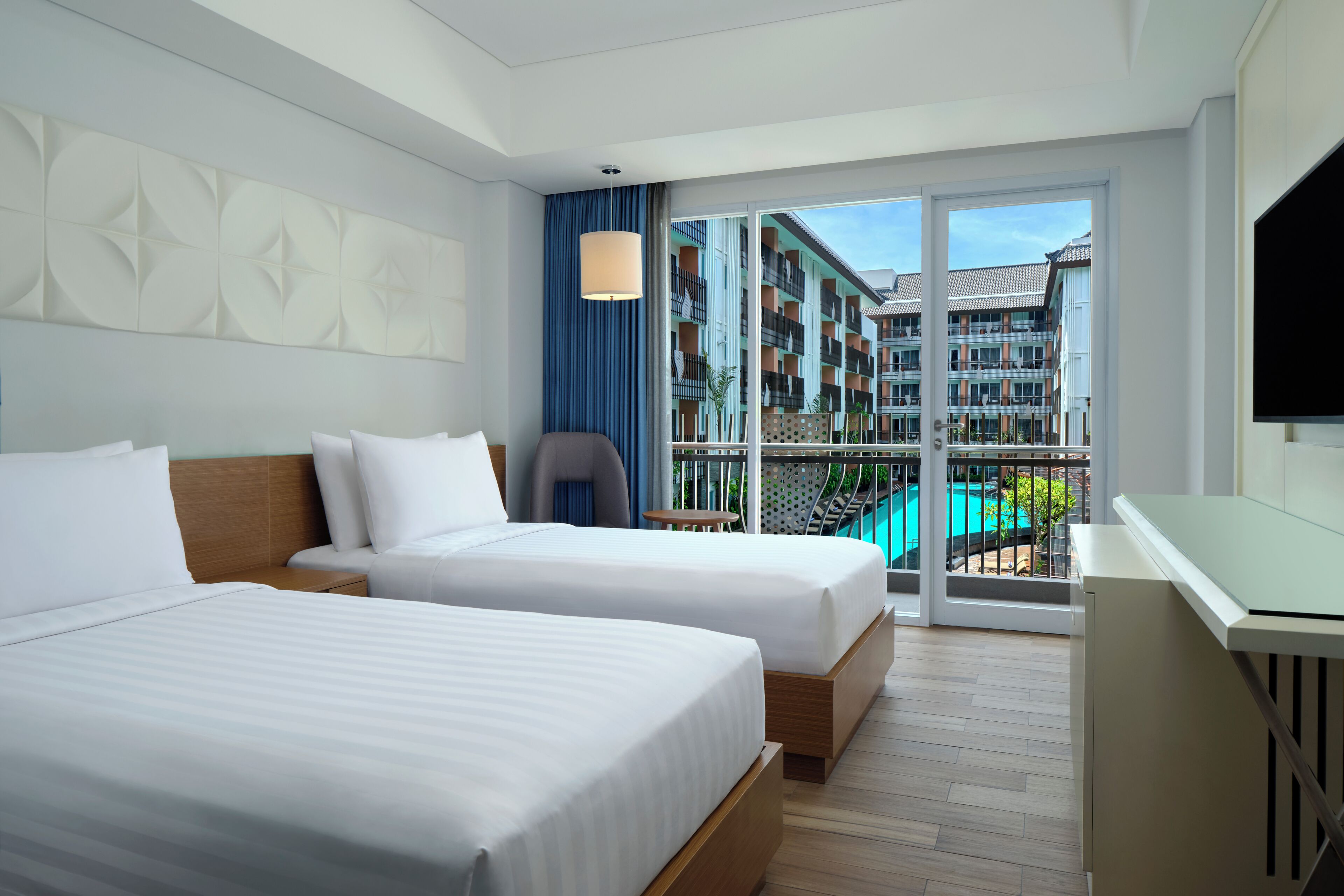 A hotel room with two beds, a TV, a balcony with a pool view, and a cityscape in the background.