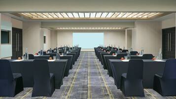 A conference room with rows of tables, chairs, and water bottles, equipped with a large screen at the front.