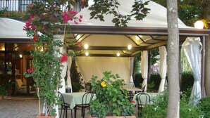 Un patio al aire libre con mesas y sillas, rodeado de plantas en macetas y una zona de estar cubierta.