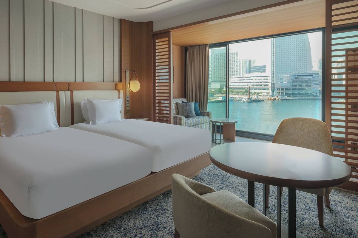 A hotel room with two beds, a small seating area, and a view of a cityscape and water.