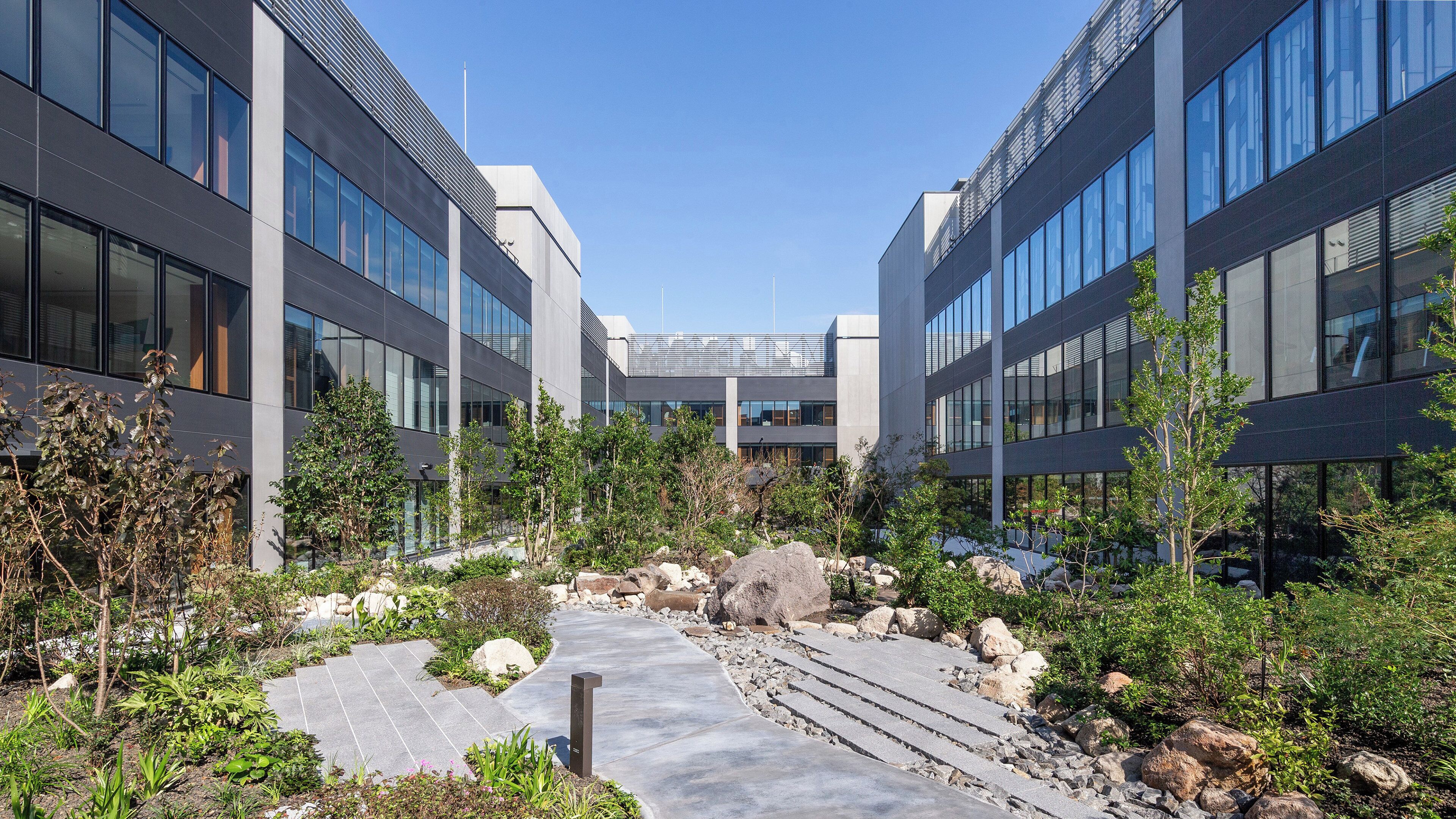 A modern building with large windows and a central courtyard with a pathway, surrounded by greenery and rocks.