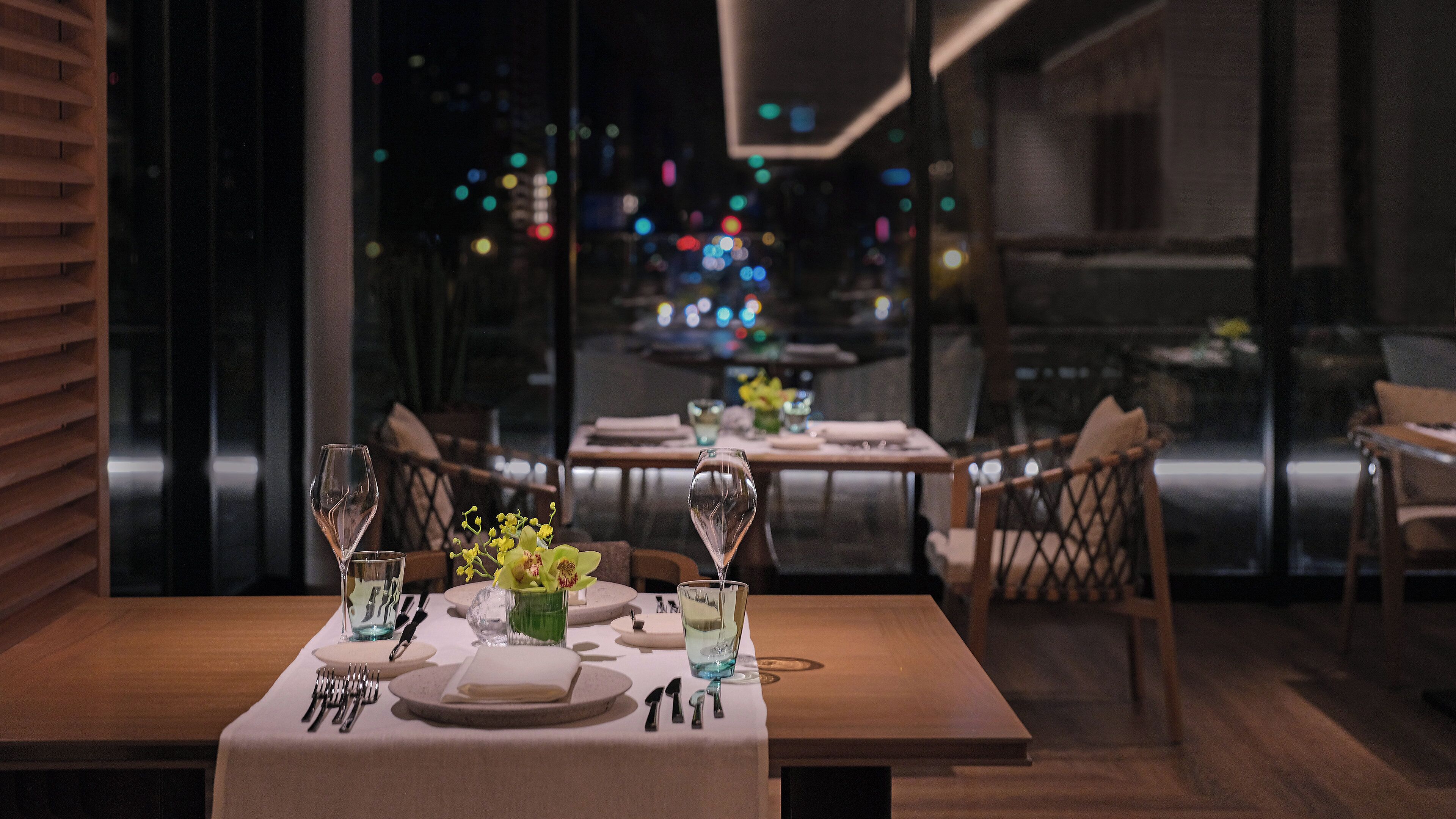 A restaurant with a table set for two, featuring a vase with flowers, a glass of water, and a wine glass. The table is positioned near a window with a view of the city at night.