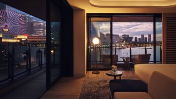 A modern hotel room with a large window offering a city view, a cozy seating area, and a small table with a lamp.