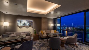 Habitación de hotel moderna con sofá, sillones, una mesita de centro y vistas a un paisaje urbano nocturno.