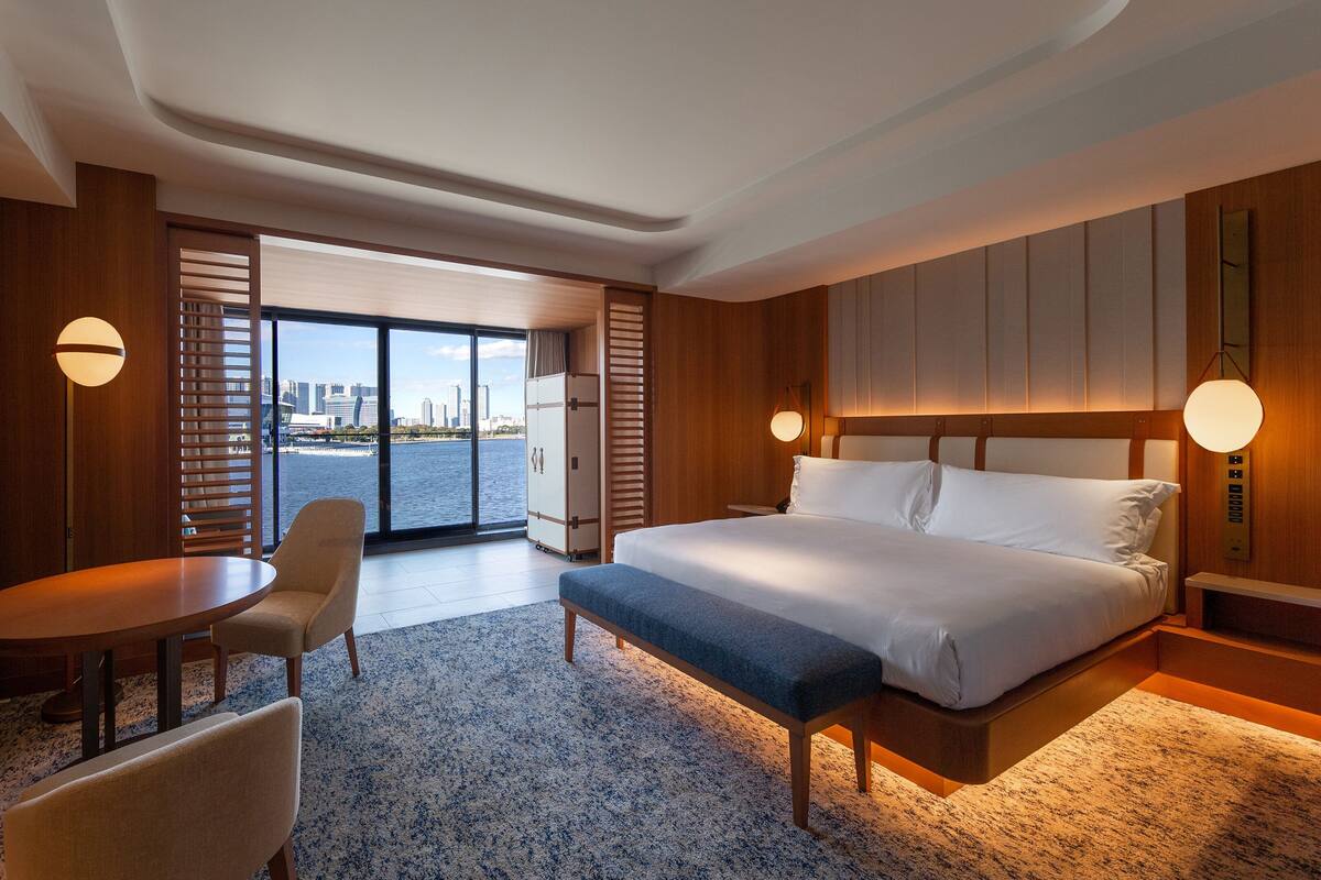 A hotel room with a bed, a desk, a chair, a TV, and a view of a city skyline through large windows.