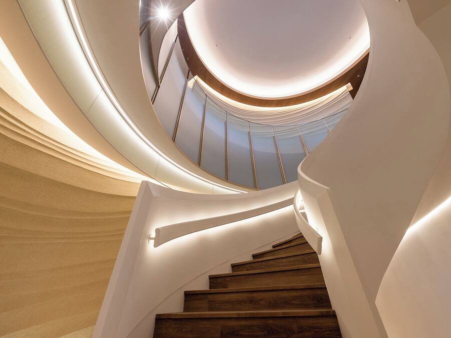 A spiral staircase with wooden steps and white walls, leading upwards to a light-filled area.