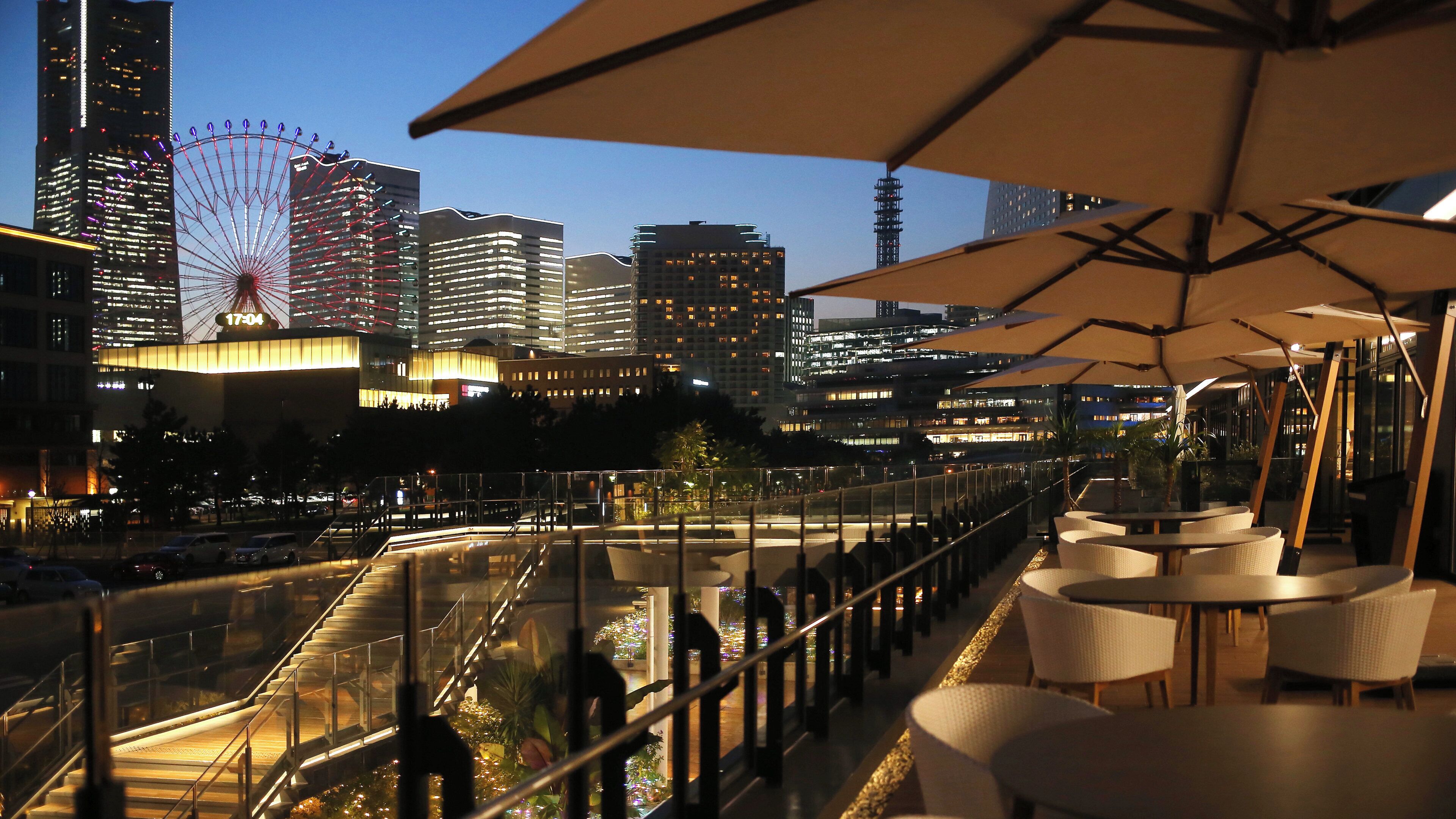 An outdoor seating area with tables and umbrellas, overlooking a cityscape with illuminated buildings and a Ferris wheel.