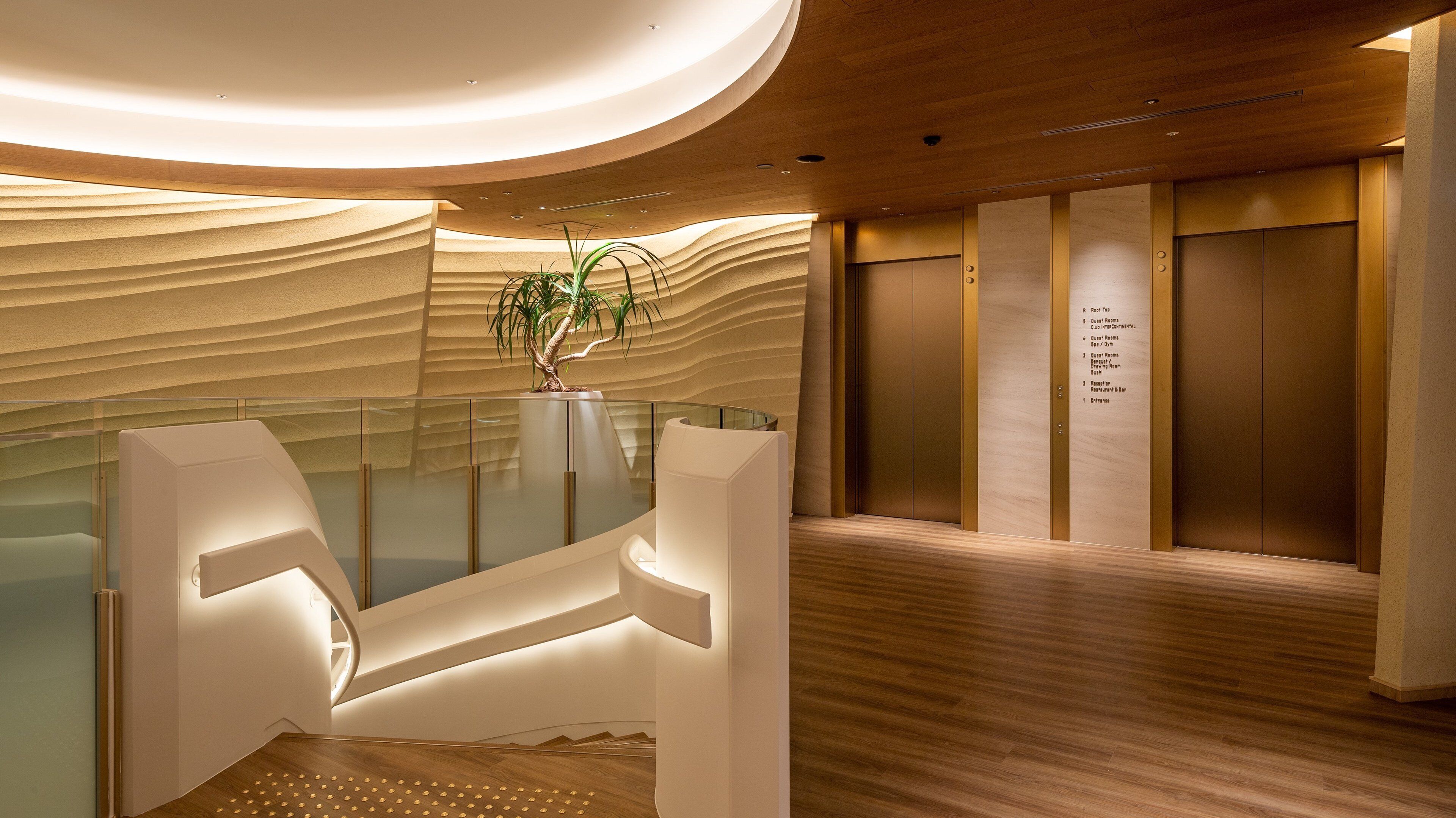 A modern interior with a reception desk, a glass partition, a potted plant, and a wall-mounted elevator control panel.