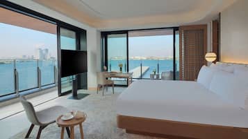 Habitación de hotel moderna con una cama grande, un escritorio, una silla y un televisor. Se ve el agua y edificios a través de la ventana.