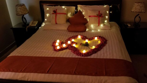 A bed with a heart-shaped arrangement of candles and rose petals, a red blanket, and pillows with red ribbons. - Luxury Inn (Karachi)